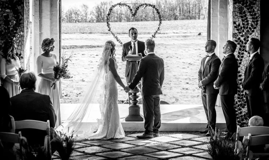A black and white photo of a bride and groom on a wedding platform standing in front of a priest and a large hanging heart made from leaves.