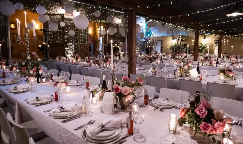 A wedding reception set up with white table cloths, flowers and fairly lights next to grey stone walls.